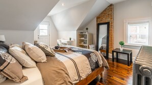 A bedroom with a sloped ceiling, a bed with multiple pillows, a small table, a radiator, and a window with blinds. - Boutique Penthouse Designer 1 BR Suite w/2 Balconies in Downtown Toronto (Toronto)