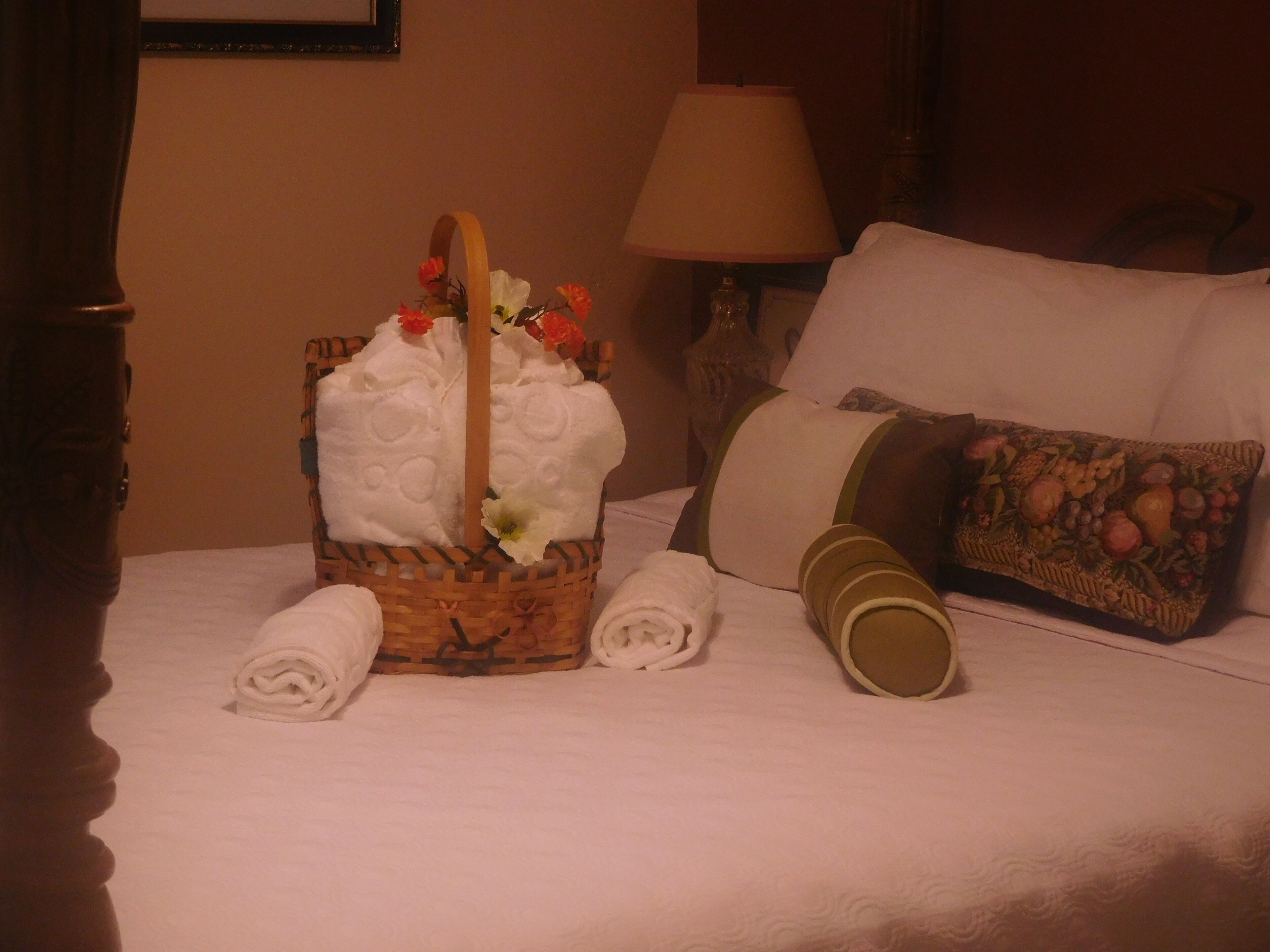 A neatly made bed with a basket of folded towels, a pillow with a fruit pattern, and a bedside lamp.