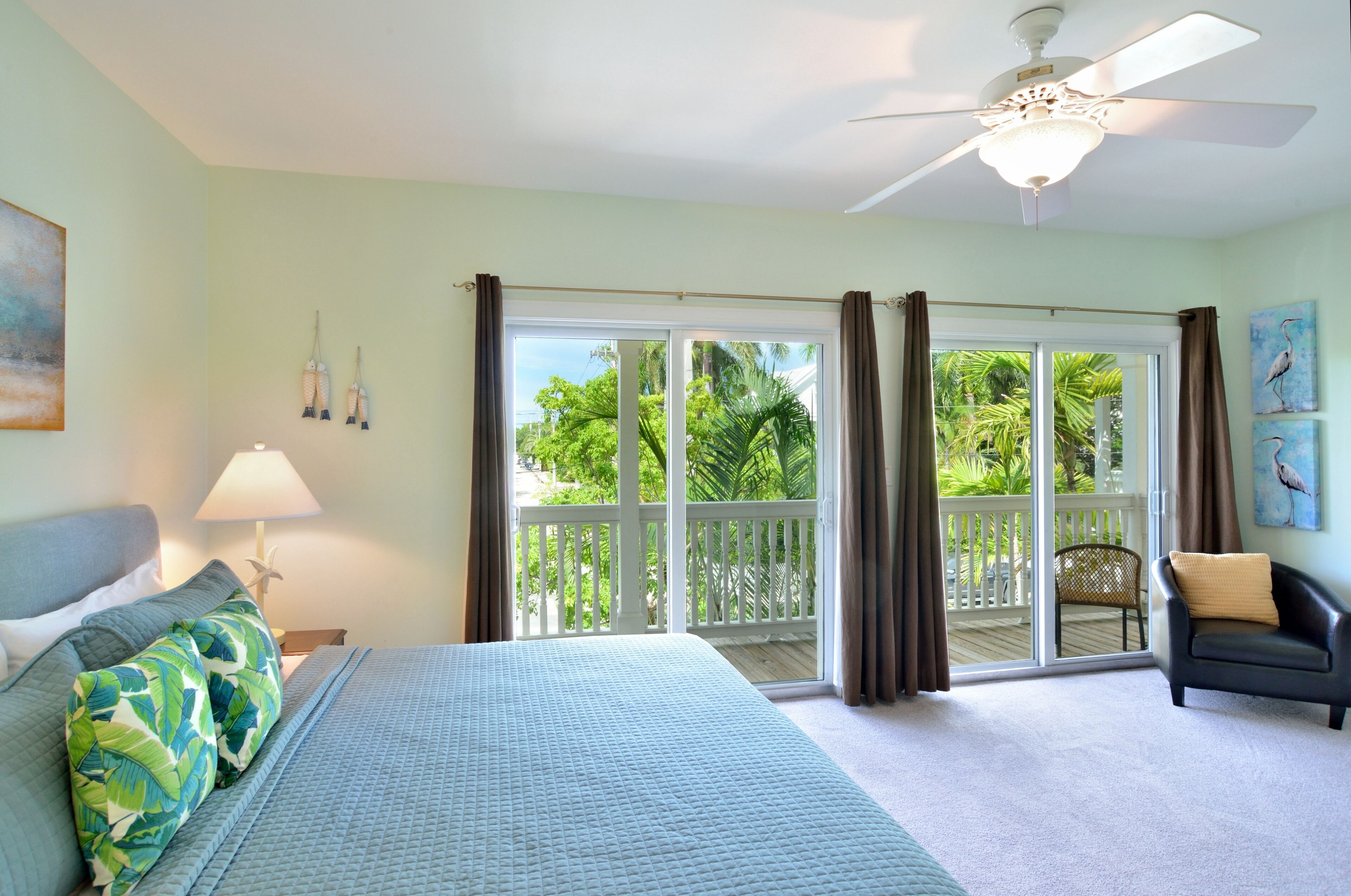 Une chambre à coucher comprenant un lit, une table de chevet, une lampe, un ventilateur de plafond et une vue sur un balcon avec une chaise.