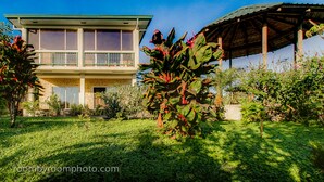 A two-story house with a balcony, surrounded by lush greenery and a small garden.