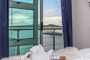 A room with a large window offering a view of a body of water, a pier, and a distant hill. - 1BR Sub-Penthouse - Panoramic Views (Auckland)