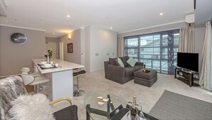 A modern living room with a sofa, ottoman, and a glass coffee table. There is a TV and a large window with curtains. - 1BR Sub-Penthouse - Panoramic Views (Auckland)