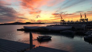 Un puerto al atardecer con barcos, grúas y un edificio grande con techo curvo.