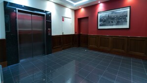 Un hall d’entrée avec un ascenseur noir, un mur rouge orné d’un tableau encadré et un sol carrelé.