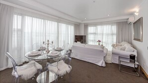 A modern hotel room with a glass dining table, white furniture, and large windows with sheer curtains. - Panoramic Views - 2 Bedroom Sub-Penthouse (Auckland)