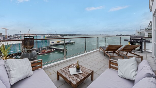 A rooftop terrace with a view of the marina, including boats and cranes, furnished with wooden chairs and a table with a bottle and glasses. - Panoramic Views - 2 Bedroom Sub-Penthouse (Auckland)