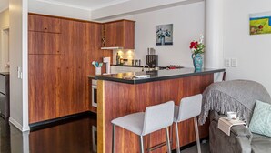A modern kitchen with wooden cabinets, a central island with bar stools, and a dining area with a sofa and a coffee table. - Princes Wharf - 1BR Comfortable Luxury (Auckland)