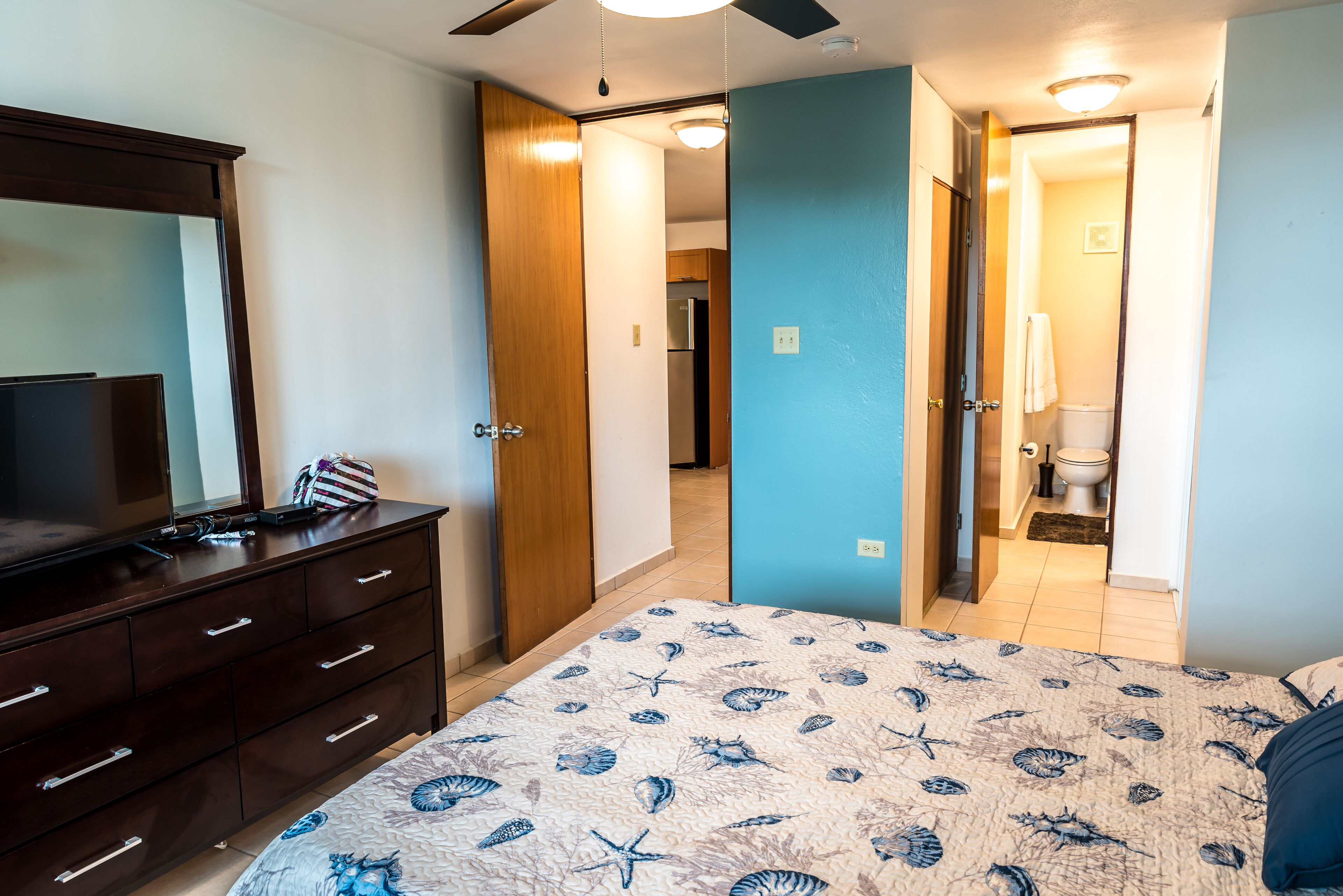 Une chambre à coucher comprenant un lit, une commode et un téléviseur. Des portes donnent accès aux autres pièces.