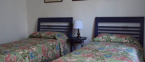A room with two single beds, each with floral bedding, a small bedside table with a lamp, and three framed pictures on the wall.
