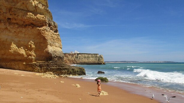 Ein Sandstrand mit einer Klippe, Wellen im Meer und einer Person, die spazieren geht.