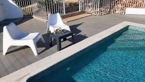 Ein Pool mit einer gefliesten Kante, zwei weiße Liegestühle und ein kleiner Tisch mit einem Glas und Blumen.