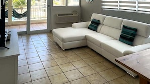 Ein Wohnzimmer mit einem weißen Sofa, einem kleinen Tisch, einem Fernseher und Blick ins Freie durch große Fenster.