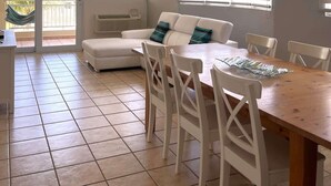 Ein Essbereich mit Holztisch und weißen Stühlen, eine weiße Couch mit Türkis-Kissen, einer dekorativen Deckenleuchte und Blick durch Glastüren.