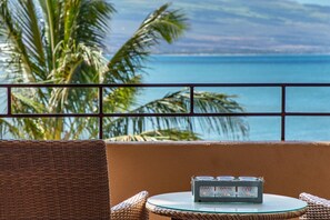 A wicker chair and table on a balcony with a view of the ocean and palm trees.