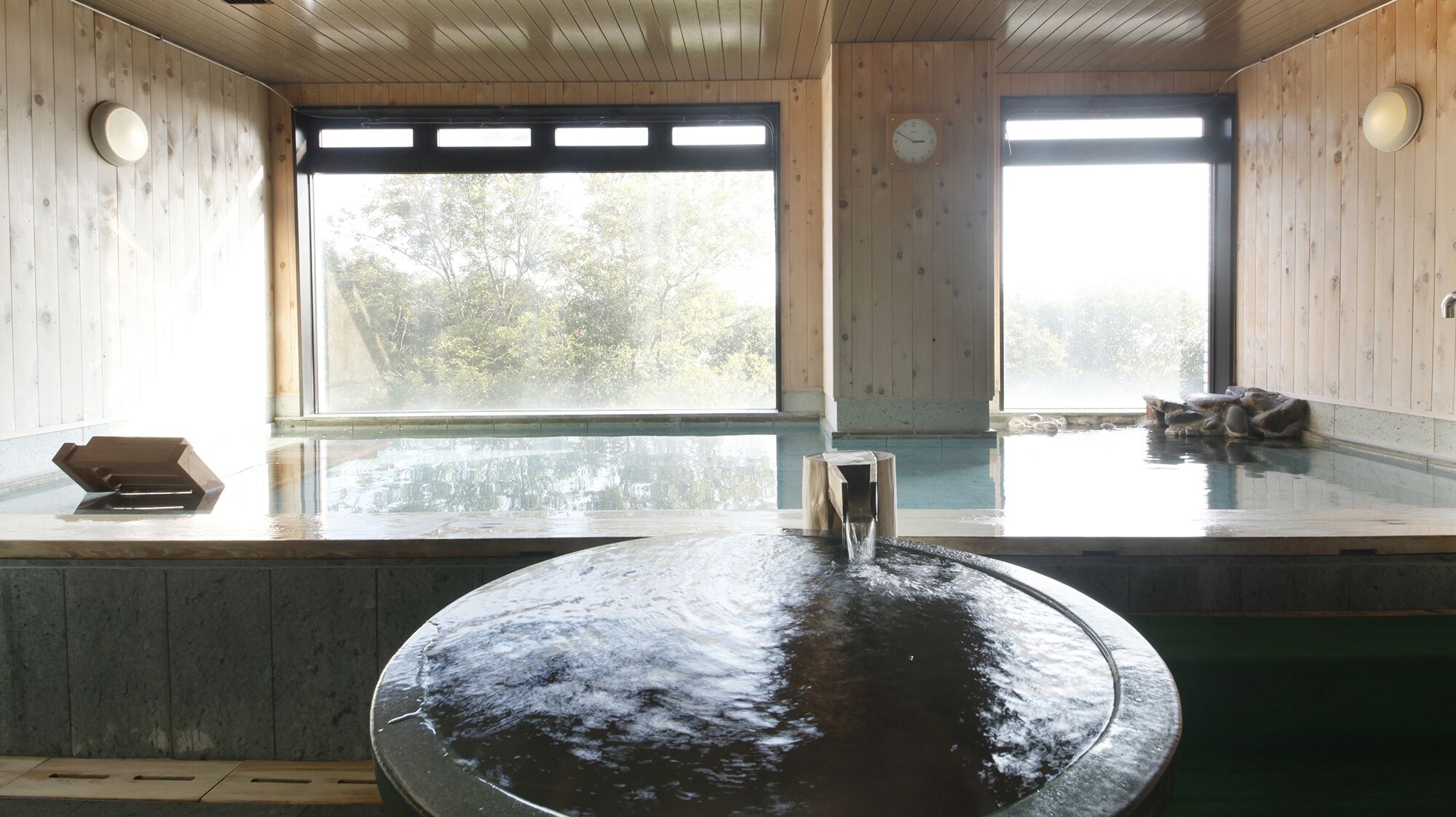 A wooden spa with a large hot tub and a smaller soaking area, large windows with a view of trees, and a wooden ceiling.
