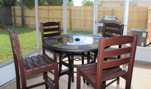 A round glass table with four wooden chairs, a barbecue grill, and a wooden fence in the background.