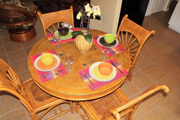 A round wooden dining table with four chairs, each with a woven backrest. The table is set with plates, bowls, and a vase with flowers.