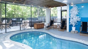 Une piscine intérieure avec vue sur l’extérieur, entourée de chaises longues et d’un espace barbecue.