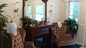 Une pièce avec une cheminée, deux fauteuils marron, un grand miroir et une fenêtre donnant sur un paysage verdoyant.