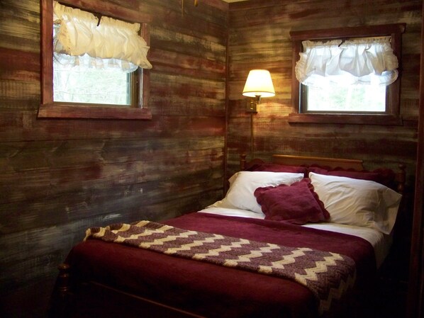 Una habitación de madera con una cama, dos ventanas con cortinas, una lámpara y una manta.