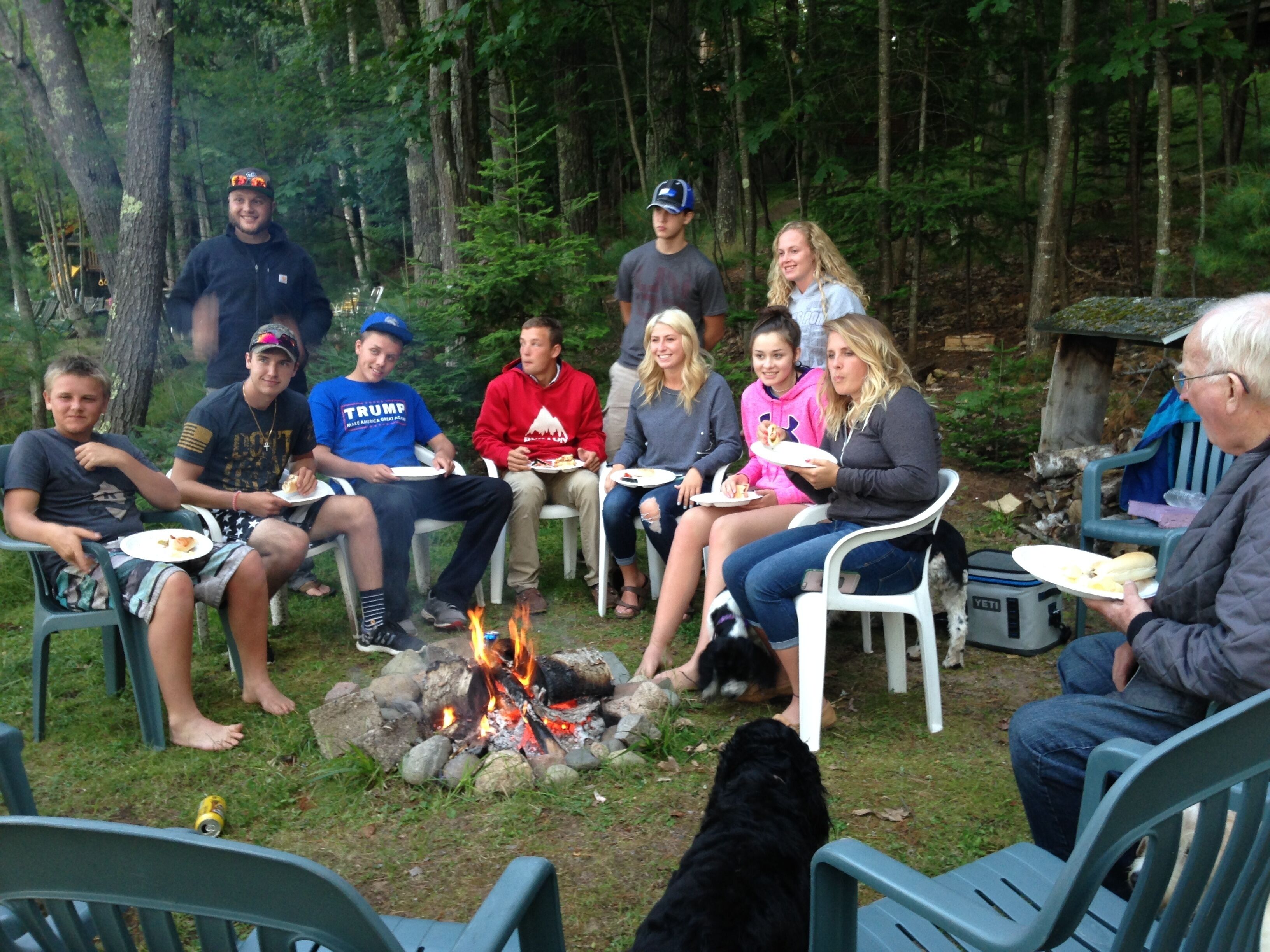 A group of people sitting around a campfire in a wooded area with plastic chairs and a cooler.