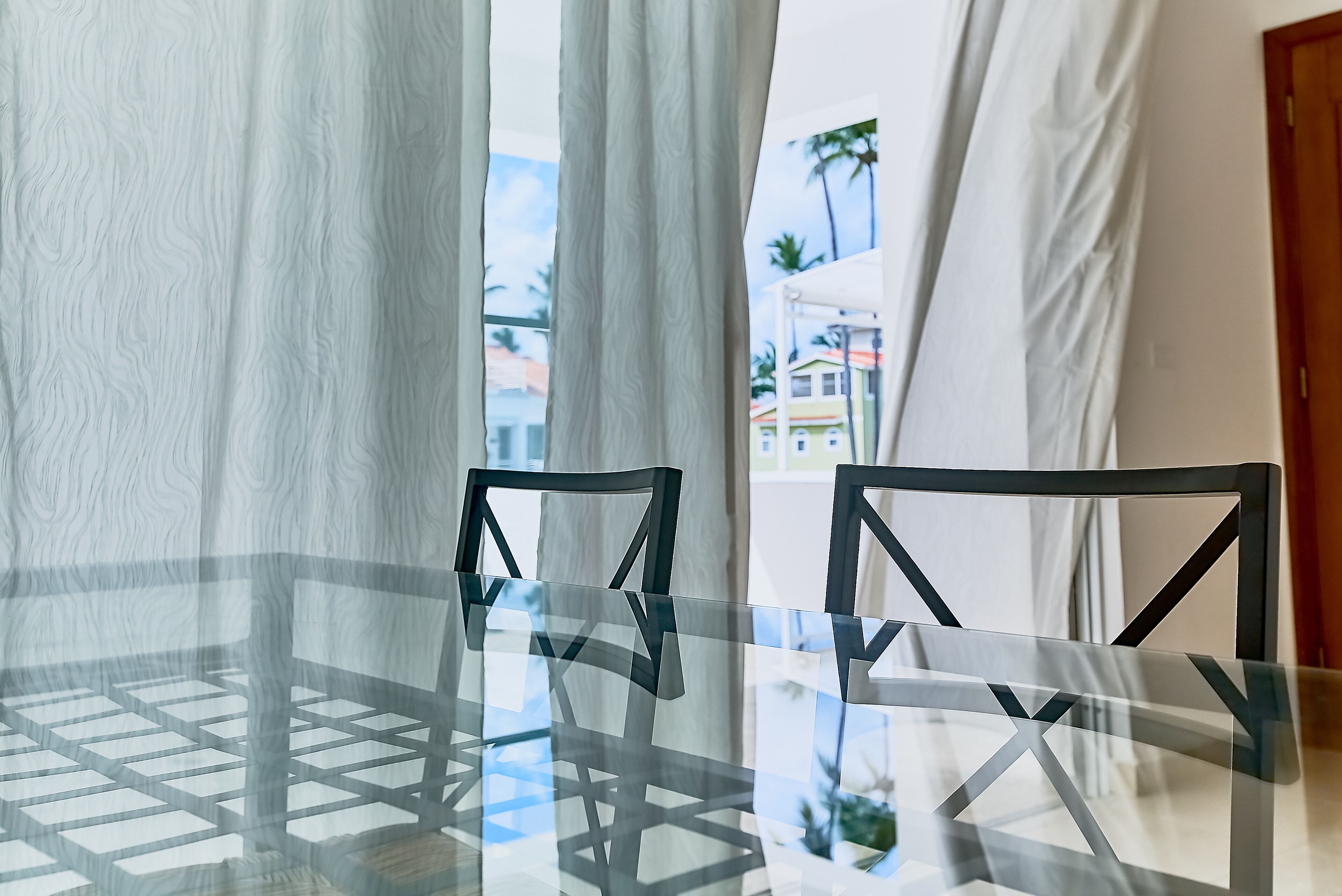 A glass-top table with two black chairs, a white curtain, and a view of buildings and palm trees outside.