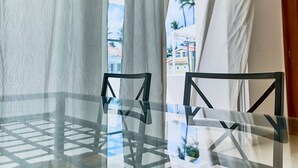 A glass-top table with two black chairs, a white curtain, and a view of buildings and palm trees outside. - Los Corales Beach Village - Your private paradise - 2BR first floor - LT#B2 (Punta Cana)
