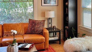 A living room with a brown leather sofa, a red patterned rug, a coffee table with a vase and remote control, a black bookshelf, and a window with blinds. - Titania - Second Spring Property 1-3 Bedroom Luxury Rentals In Downtown Ashland (Ashland)