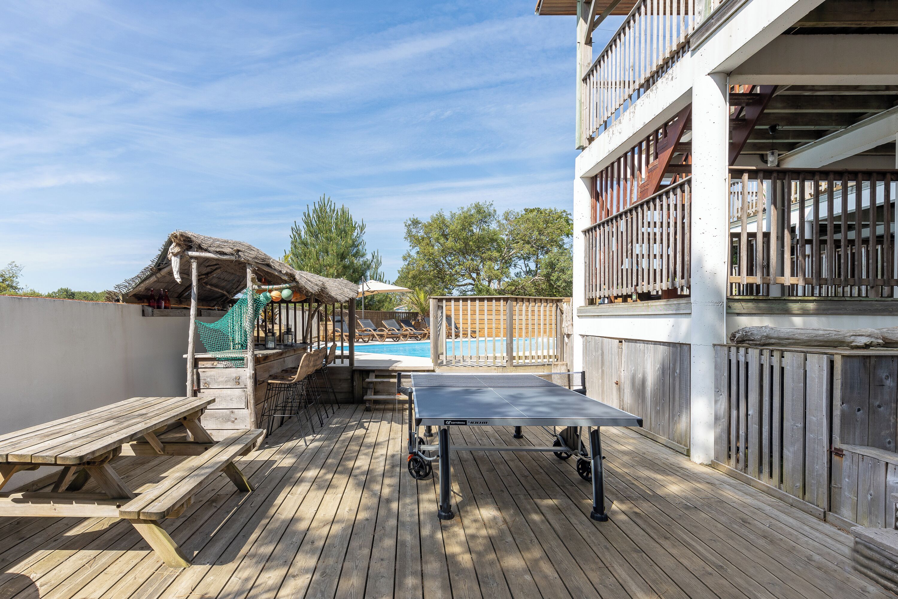 Une terrasse en bois avec une table de ping-pong, une piscine et une structure recouverte d’un toit de chaume.