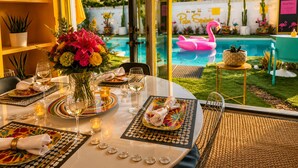 Un coin repas avec une table dressée pour deux personnes, un vase de fleurs et une vue sur une piscine agrémentée d’un flotteur en forme de flamant rose.