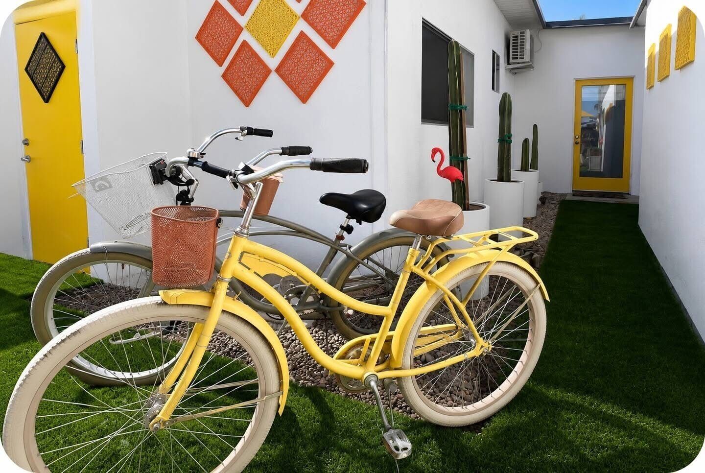 A yellow bicycle with a basket and a seat, parked on a grassy area with a white wall and decorative elements in the background.