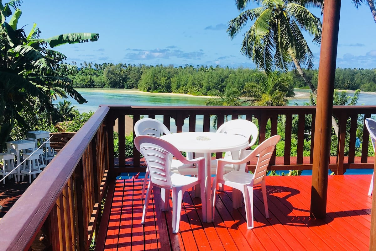 A red deck with white plastic chairs and a table, overlooking a beach and palm trees.