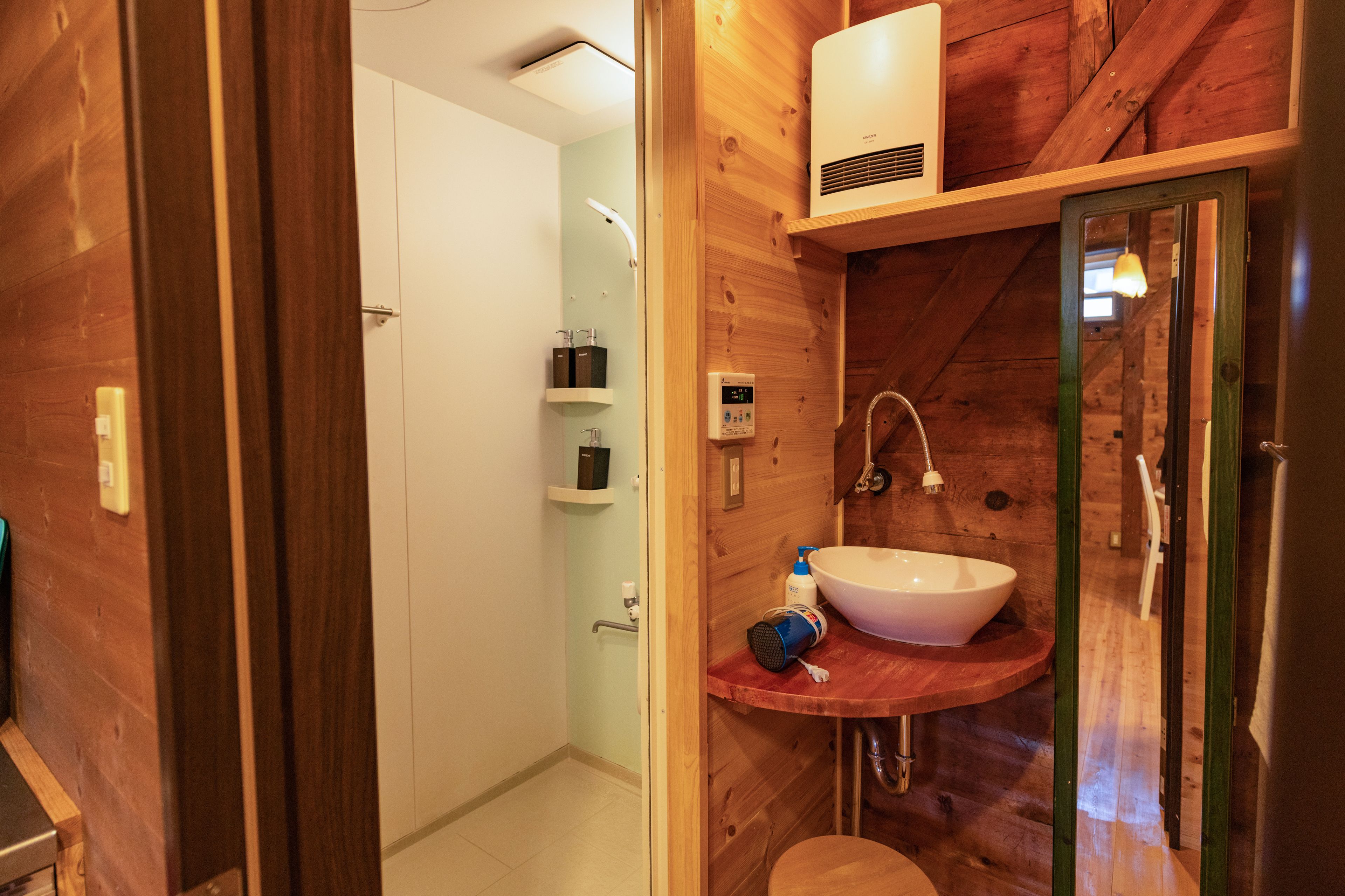 A wooden bathroom with a white sink, a shower, and a toilet. There is a shelf with toiletries and a towel rack.