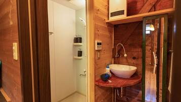 A wooden bathroom with a white sink, a shower, and a toilet. There is a shelf with toiletries and a towel rack.