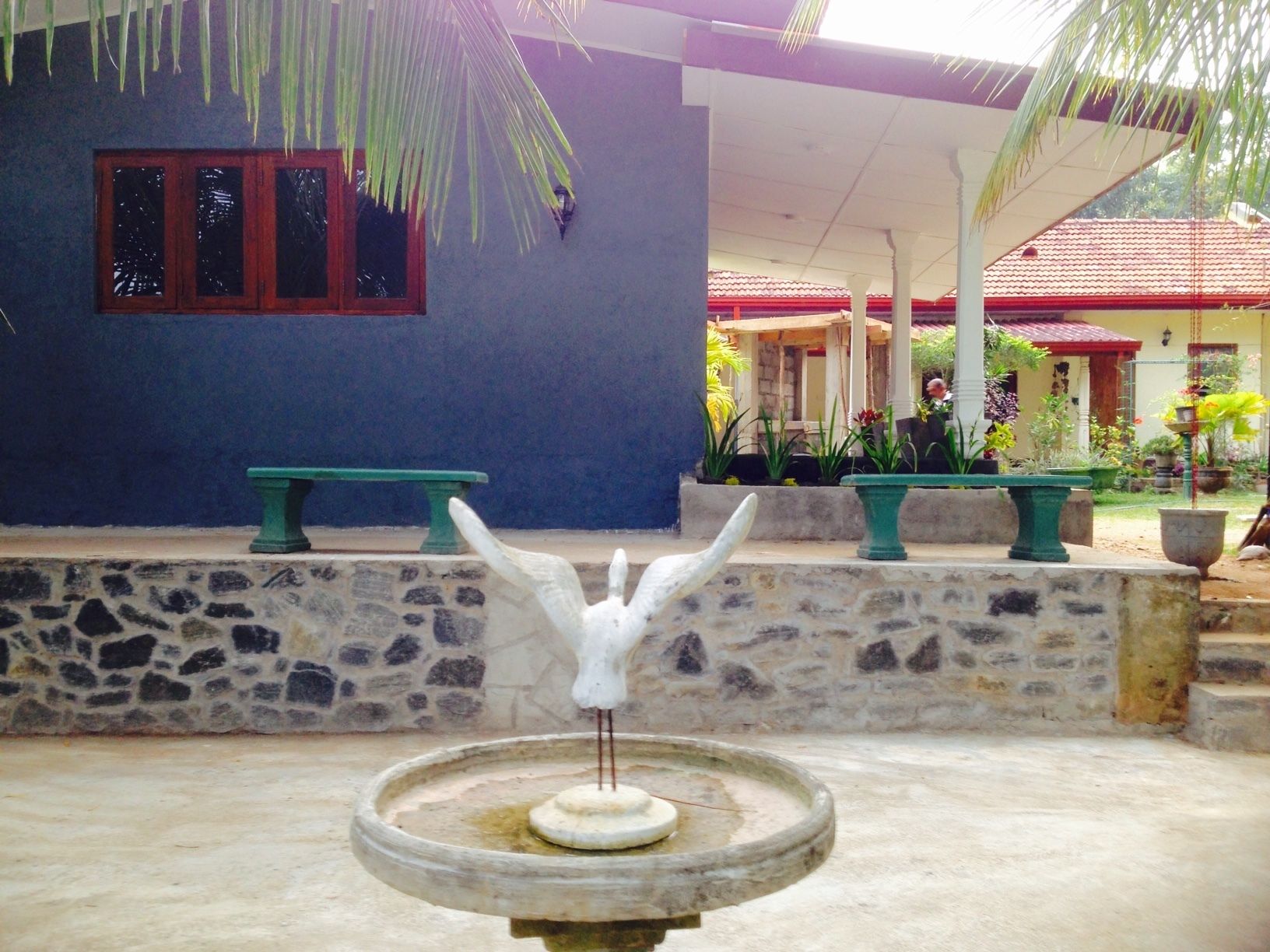 Une fontaine en pierre ornée d’une sculpture blanche au centre, placée devant un mur bleu agrémenté d’une fenêtre et d’un banc.
