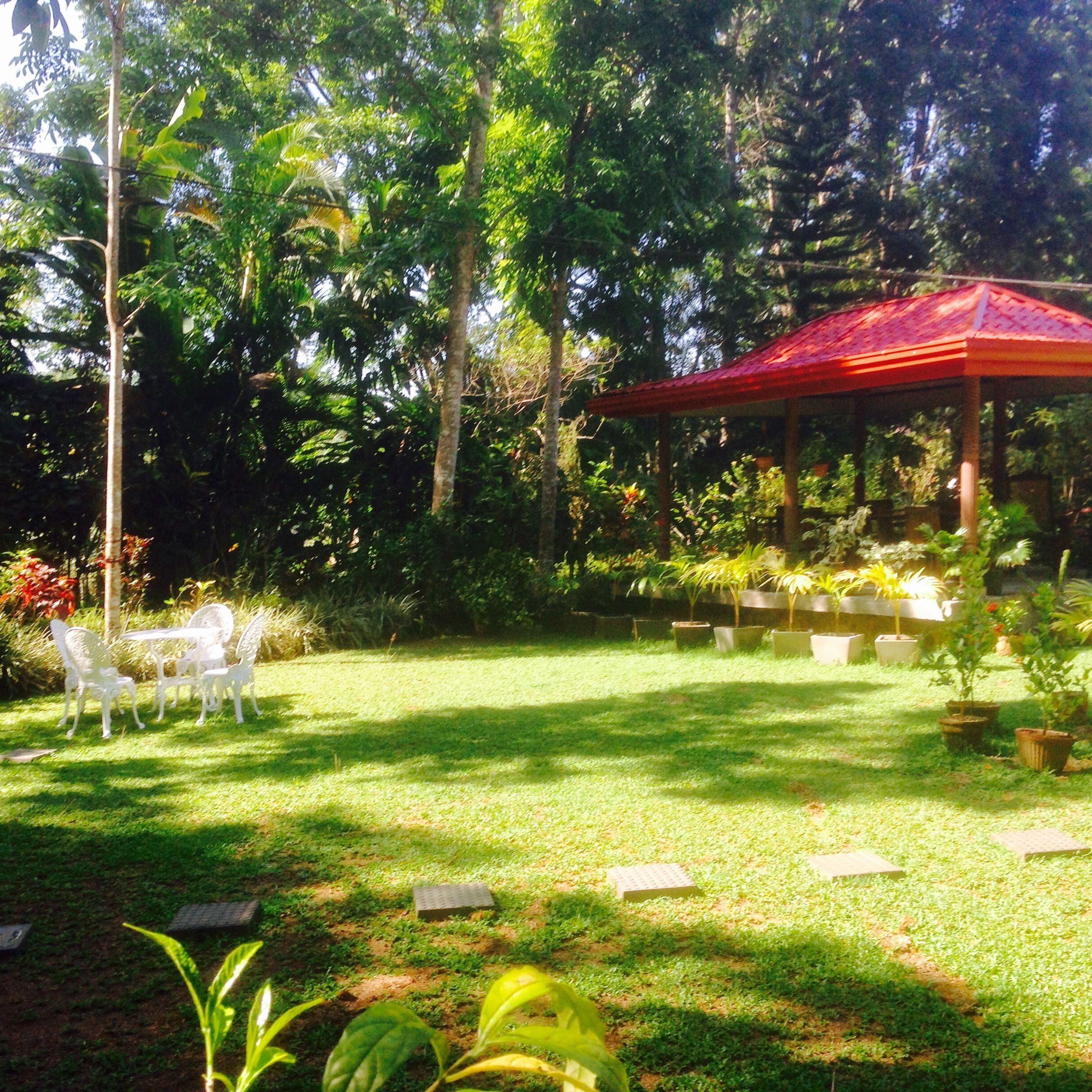 Um jardim com um gazebo vermelho, cadeiras brancas e plantas em vasos.
