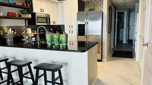 A modern kitchen with a black countertop, stainless steel appliances, white cabinets, and a bar stool area. - ROMANTIC MALIBU ELEGANCE - on HGTV & DIRECTLY on the LUMINOUS GULF SAND! (Orange Beach)