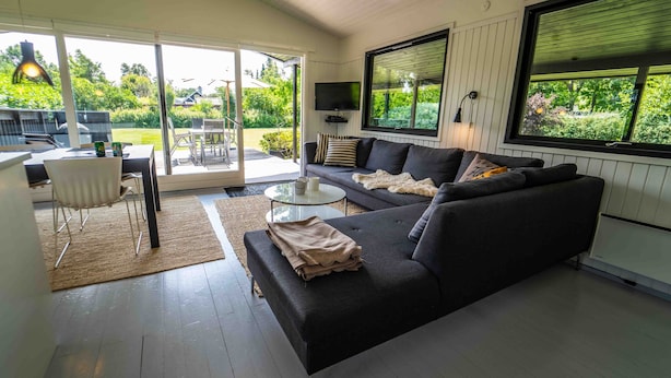 A modern living room with a dark sofa, a glass coffee table, and a dining area with a white chair and table. - Cottage 150 m from Beach (Stege)