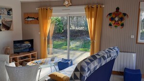 Un salon comprenant un téléviseur, un canapé et une table, une porte-fenêtre coulissante donnant sur un jardin, et un mur orné d’une fresque représentant des poissons.