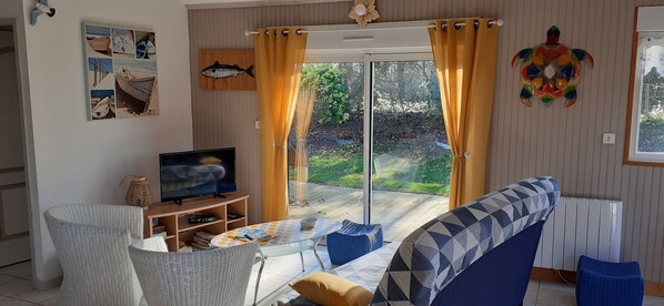 Ein Wohnzimmer mit Fernseher, einer Couch und einem Tisch, einer Schiebetür zu einem Garten sowie einer Wand mit einem Fischfries.