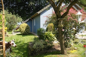 Ein Haus mit blauer Fassade, einer Holzleiter und ein Garten mit verschiedenen Pflanzen und einem Baum mit rosa Blüten.