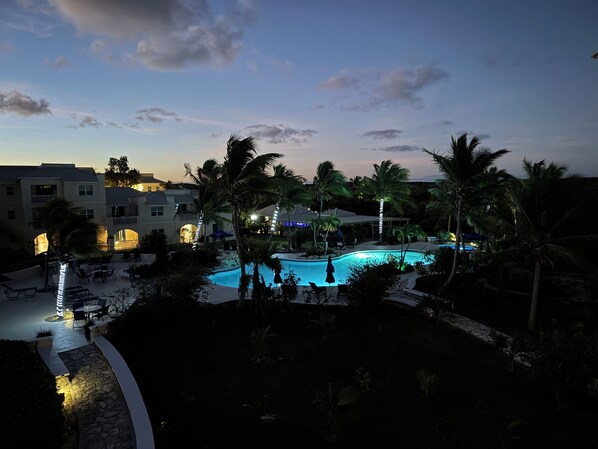 Zona de piscina en un resort al anochecer, con palmeras, sillas de descanso y un edificio con ventanas iluminadas.