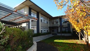 Un edificio moderno con una galería techada, un jardín ajardinado y un árbol con hojas otoñales.