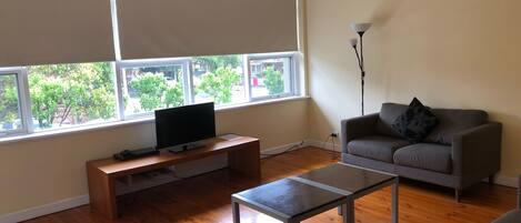 A living room with a TV on a wooden stand, a grey sofa, a floor lamp, and a window with blinds.