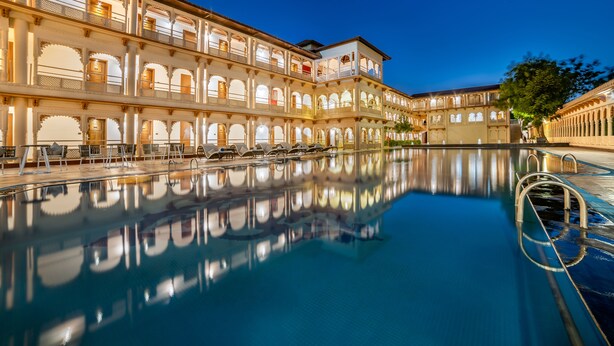 A large, well-lit swimming pool with clear reflections, surrounded by a multi-story building with balconies and arched windows. - Gateway Kandla Gandhidham (Anjar)