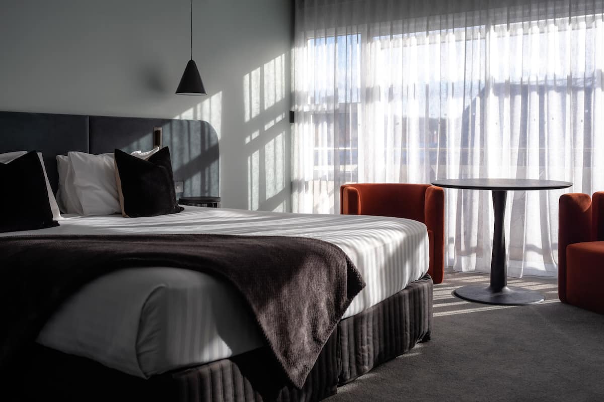 A modern hotel room with a large bed, a round table, and a view through sheer curtains.