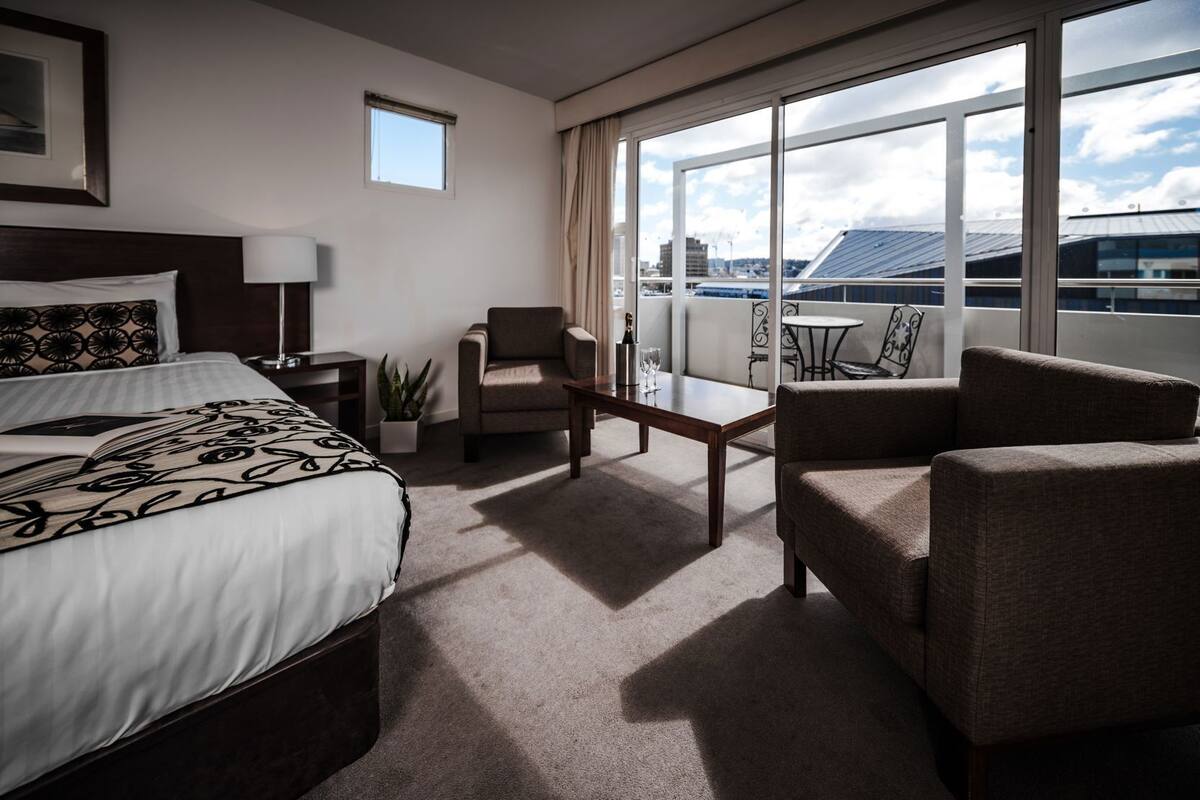A hotel room with a bed, a chair, a small table, a plant, and a view of the city through large windows.