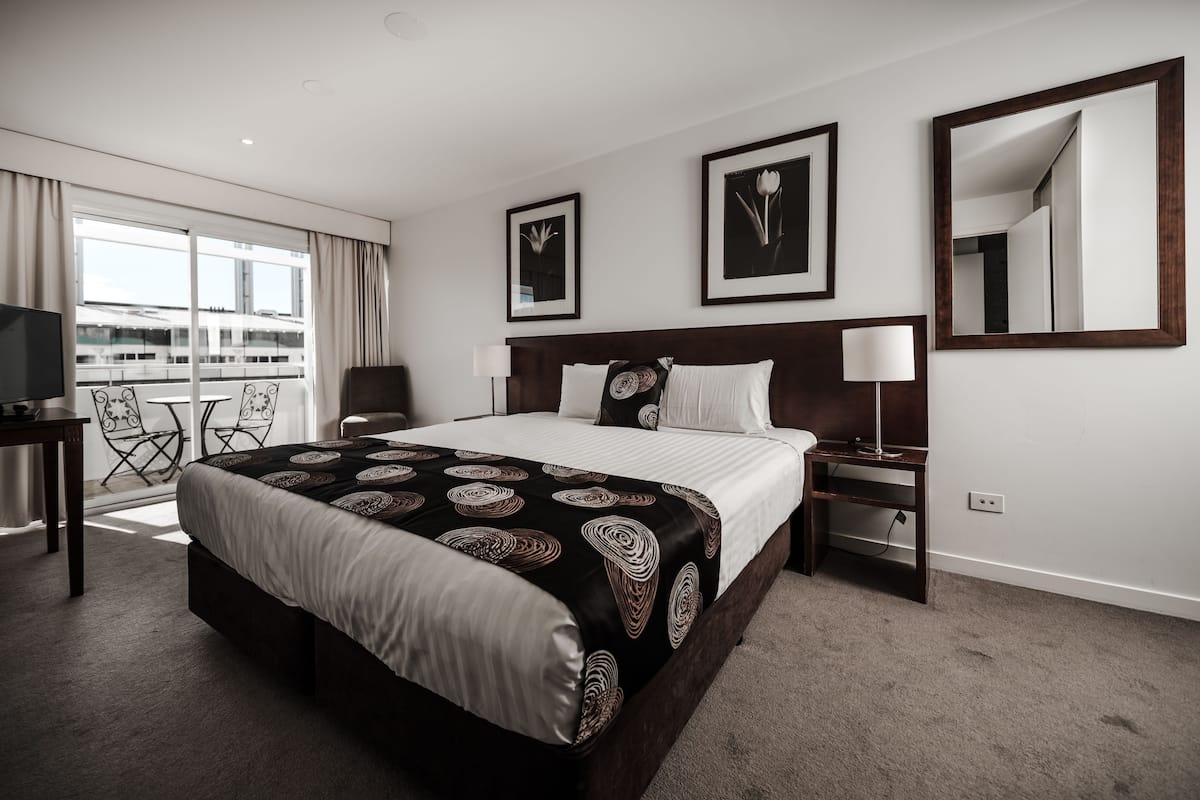 A hotel room with a large bed, a desk with a chair, a TV, a balcony with a view, and a mirror on the wall.
