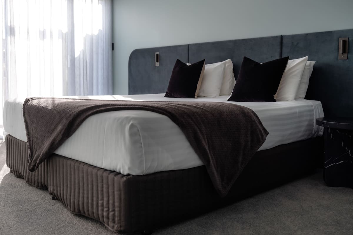A modern hotel room with a large bed, dark headboard, and a view of the city through sheer curtains.