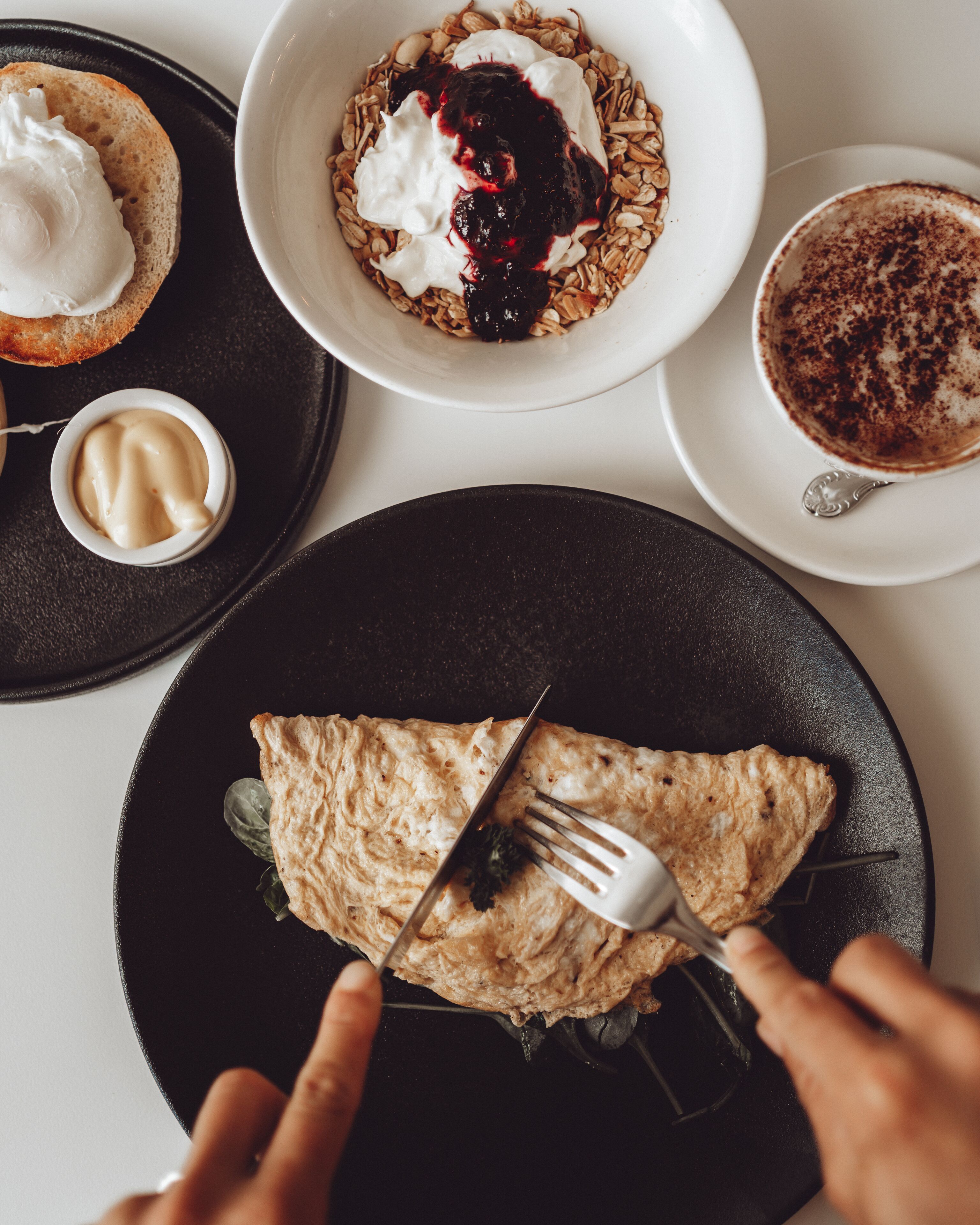 Una escena de desayuno con crepas en un plato, granola con yogur y frutos rojos en un tazón, una taza de café y un recipiente pequeño de mantequilla.